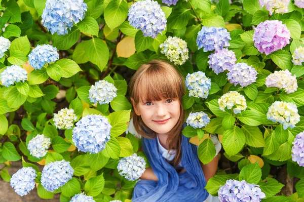 Outdoor portrait of adorable little girl of 6-7 years old, sitting between beautiful blue hydrangea flowers, covering with purple scarf — Stock Photo, Image
