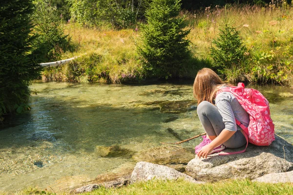 Cute little girl of 7-8 years old hiking in swiss Alps, resting by the river, wearing sport clothes, trainers and backpack, back view — Stock Photo, Image