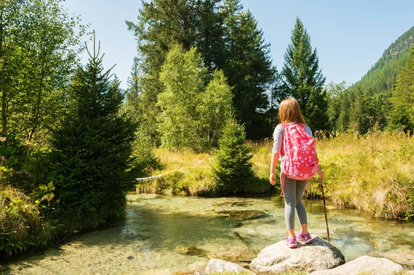 Gadis kecil lucu berusia 7-8 tahun hiking di Alpen swiss, beristirahat di tepi sungai, mengenakan pakaian olahraga, pelatih dan ransel, melihat ke belakang — Stok Foto