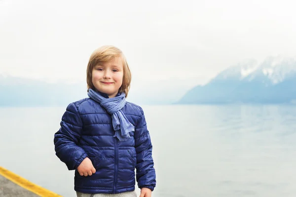 Retrato ao ar livre de adorável menino loiro de 4-5 anos, vestindo casaco azul e cachecol, em pé junto ao lago em um dia nublado — Fotografia de Stock