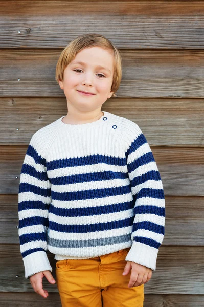 Retrato vertical de moda de niño adorable de 4-5 años con jersey blanco cálido con rayas azules y pantalones amarillos, de pie sobre fondo de madera — Foto de Stock