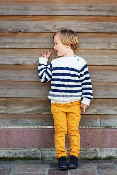 Moda pionowa portret uroczego małego chłopca z 4-5 lat ubranego w ciepły biały sweter z niebieskimi paskami i żółtymi spodniami, stojąc na drewnianym tle — Zdjęcie stockowe