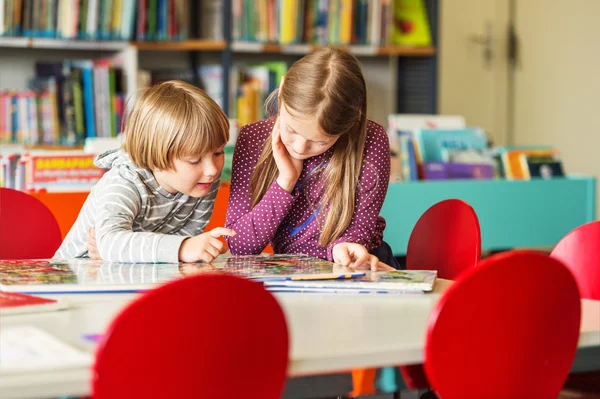 Two adorable kids, little girl and her brothe reading book in a library — Stock Photo, Image