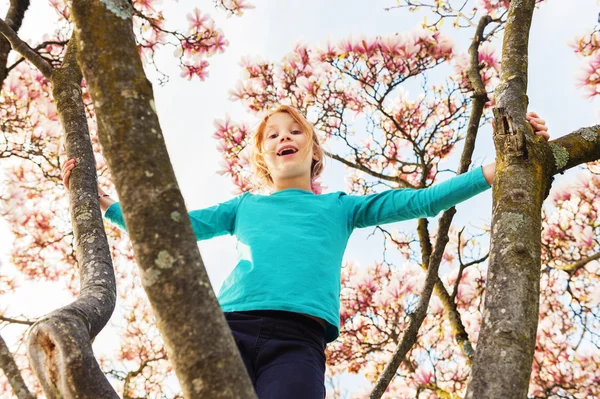 Очаровательная рыжая девочка играет на дереве магнолии в солнечный весенний день в голубой рубашке. — стоковое фото
