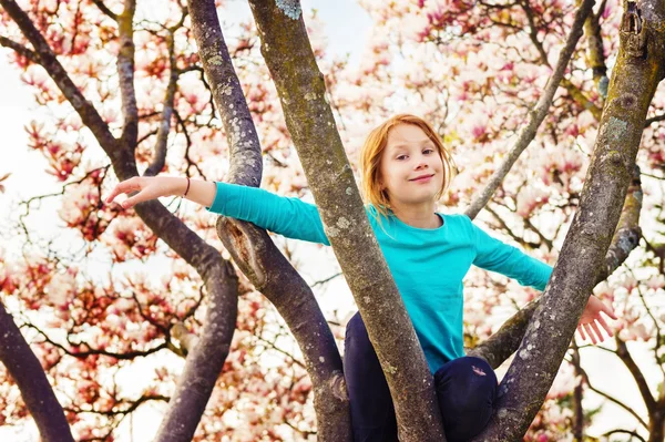 Entzückendes kleines rothaariges Mädchen, das an einem schönen sonnigen Frühlingstag auf einem Magnolienbaum spielt und blaues Hemd trägt — Stockfoto