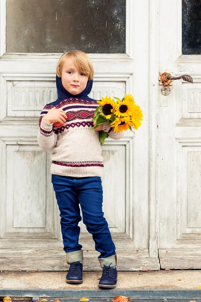 Autumn portrait of adorable little blond boy of 4 years old, wearing warm pullover with the hood, dark denim jeans and blue shoes, holding an apple and bouquet of yellow sunflowers — Stock Photo, Image