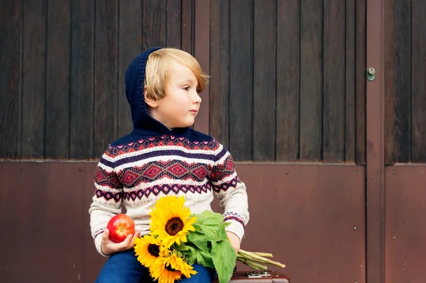 Autumn portrait of adorable little blond boy of 4 years old, wearing warm pullover with the hood, dark denim jeans and blue shoes, holding an apple and bouquet of yellow sunflowers — Stock Photo, Image