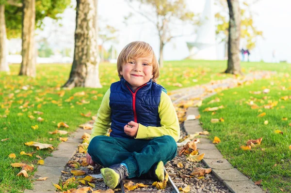 Herfst portret van een schattige kleine jongen van 4 jaar oud, spelen met gele bladeren in het park, dragen groene broek en blauw jasje — Stockfoto