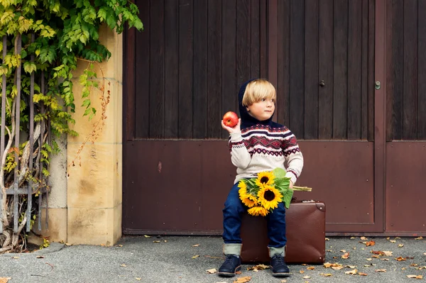 Herbstporträt eines entzückenden kleinen blonden Jungen von 4 Jahren, mit warmem Pullover mit Kapuze, dunklen Jeans und blauen Schuhen, in der Hand einen Apfel und einen Strauß gelber Sonnenblumen — Stockfoto