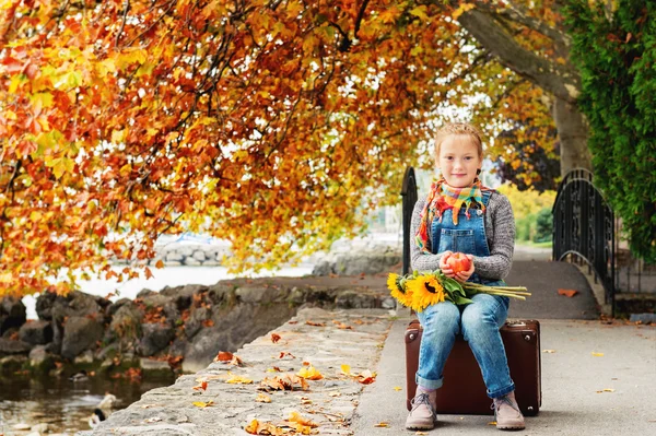 Autumn portrait of adorable little blond girl of 8 years old, wearing warm pullover, denim overalls and beige shoes, sitting on the old vintage suitcase, holding red apple and bouquet of yellow sunflowers Stock Image