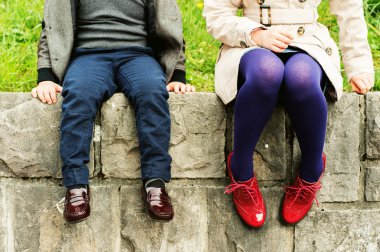 Two pairs of kids feet wearing fashion shoes, little boy wearing blue trousers and brown moccasins, schoolgirl in trench coat, purple tights and red brogues clipart