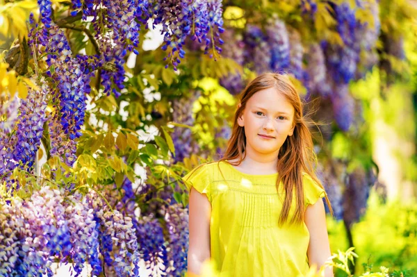 Portrait vertical élégant en plein air d'une jolie petite fille de 8-9 ans, debout à côté de belles fleurs de glycine pourpre, vêtue d'une blouse verte, bras croisés — Photo