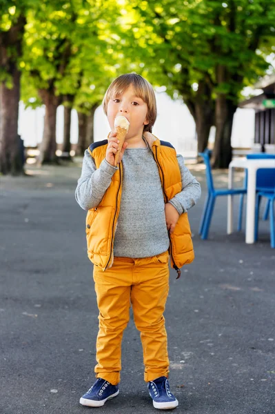 Retrato vertical al aire libre de adorable niño de 4-5 años, con jersey azul, pantalones amarillos y chaleco, comiendo helado en el parque — Foto de Stock