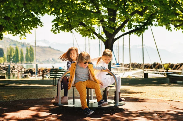 Tre barn har roligt på lekplatsen. Snygga barn spelar på karusell i parken på en mycket solig dag. Bedårande barn vänner spenderar tid tillsammans — Stockfoto