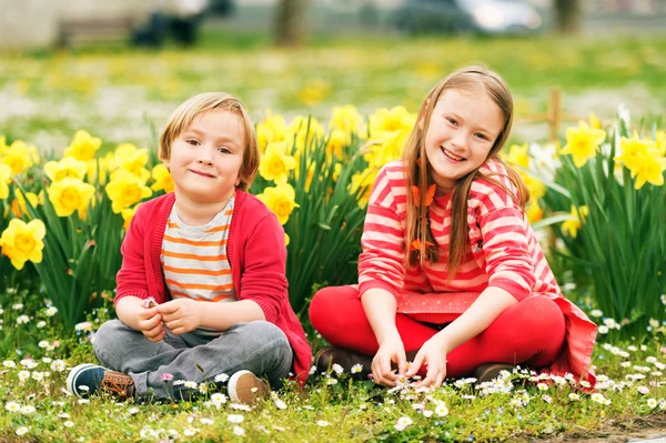 Двое милых детей, маленький мальчик и его старшая сестра, играют в парке между желтыми цветами нарциссов, одетые в ярко-красную одежду — стоковое фото