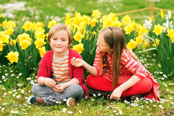 Dois meninos bonitos, menino e sua irmã mais velha, brincando no parque entre flores amarelas narcisos, vestindo roupas vermelhas brilhantes — Fotografia de Stock