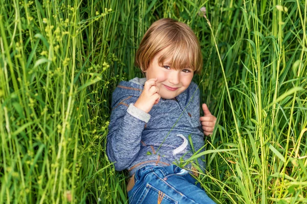 Szczery portret ładny mały chłopiec 4-5 lat, na sobie niebieski z kapturem, bawiąc się samodzielnie na zewnątrz, leżąc na trawie — Zdjęcie stockowe