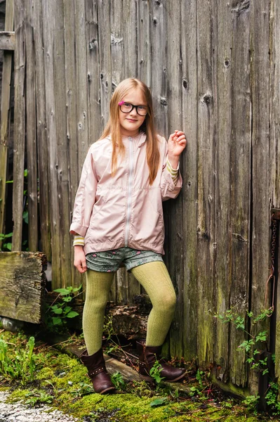 Retrato ao ar livre de uma menina bonito em óculos, de pé contra o velho fundo de madeira, vestindo meias verdes, casaco de chuva rosa pálido e botas marrons — Fotografia de Stock