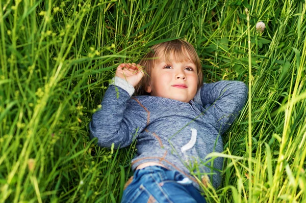 Candido ritratto di adorabile bambino di 4-5 anni, con cappuccio blu, che gioca da solo all'aperto, sdraiato sull'erba — Foto Stock