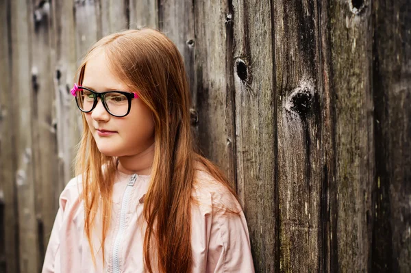 Außenporträt eines niedlichen kleinen Mädchens mit Brille, das vor altem Holzhintergrund steht — Stockfoto