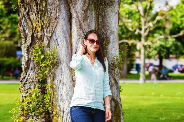 Stylische, fröhliche Brünette lehnt an einem sonnigen Tag an einem Baum in einem Park, trägt Jeans, Minzbluse — Stockfoto