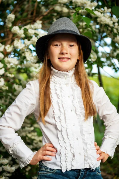 Portrait of pretty little girl of 8-9 years old in garden. Kid girl wearing white vintage blouse and grey black hat