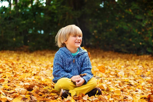 Herfst portret van een schattige kleine jongen van 4 jaar oud, spelen met gele bladeren in het park, blauwe jas en een gele broek dragen — Stockfoto