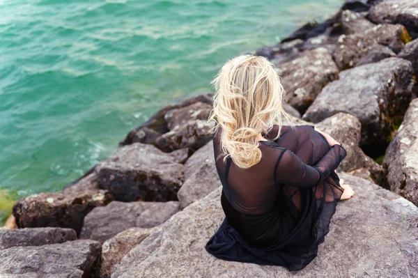 Mujer rubia romántica descansando junto al lago vistiendo vestido negro, vista trasera — Foto de Stock