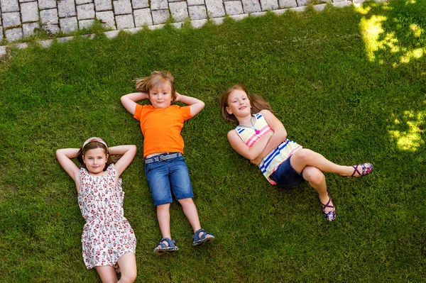 Счастливые дети веселятся на открытом воздухе. Дети играют в летнем парке. Маленький мальчик и две девочки лежат на зеленой свежей траве — стоковое фото