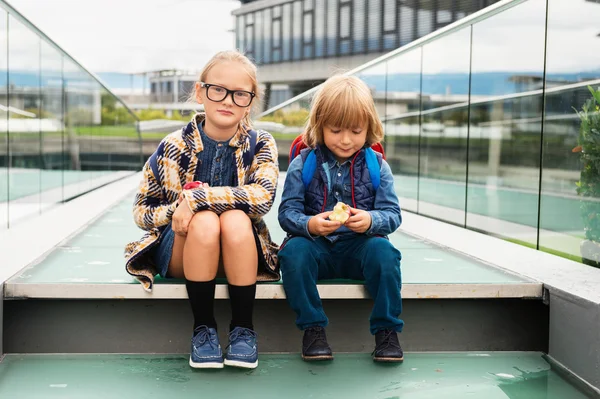 Cute kids with backpacks sitting on stairs next to school, wearing backpack, eating apples — Stock Photo, Image