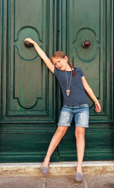 Fashion portrait of a cute little girl, wearing blue stripe t-shirt, denim shorts and grey shoes, standing next to old green door — Stock Photo, Image