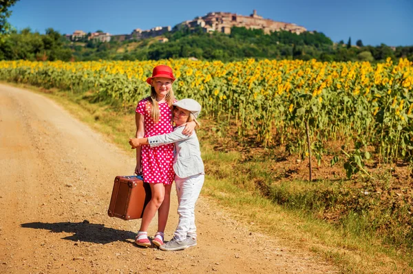Zwei niedliche Kinder, kleines Mädchen und Bruder, die die kleine Straße am Sonnenblumenfeld entlang gehen, mit einem alten braunen Koffer. Bild aufgenommen in der Toskana, Italien. Kleines Mädchen in rotem Tupfen-Kleid und Schuhen — Stockfoto