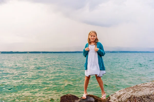 Sandalet, elbise ve mavi örgü ceket giyen 8-9 yaşında göl kenarında oynayan sevimli küçük kız — Stok fotoğraf