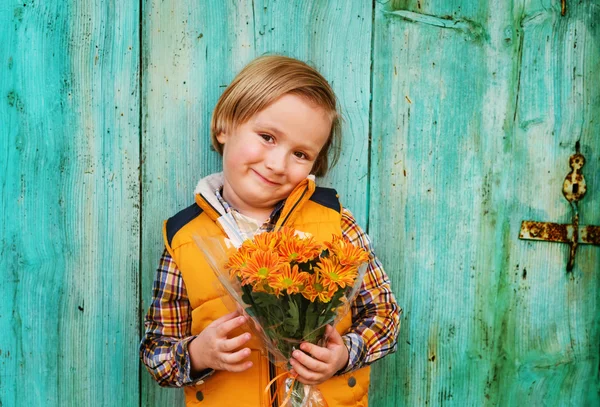 Autumn portrait of adorable little blond boy of 4 years old, wearing warm yellow vest coat, holding small bouquet of orange chrysanthemum flowers, standing in front of turquoise wooden wall — Stock Photo, Image