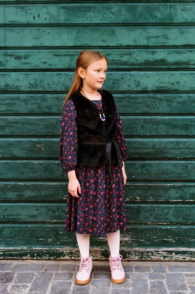 Outdoor fashion portrait of a cute little girl of 8 years old, wearing black dress and faux fur coat, standing next to green wooden wall — Stock Photo, Image