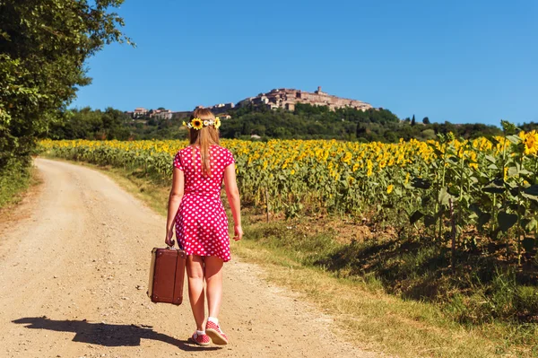 Outdoor portrait of a funny little girl walking down the road in countryside, holding old small suitcase, back view. Image taken in Tuscany, Italy