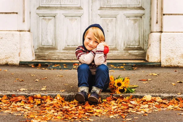 Autumn portrait of adorable little blond boy of 4 years old, wearing warm pullover with the hood, dark denim jeans and blue shoes, holding an apple and bouquet of yellow sunflowers — Stock Photo, Image