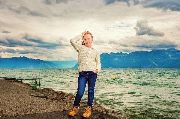 Menina bonito de 8 anos de idade jogando junto ao lago em um dia muito ventoso, vestindo pulôver de malha branca quente, braços bem abertos, imagem tonificada — Fotografia de Stock