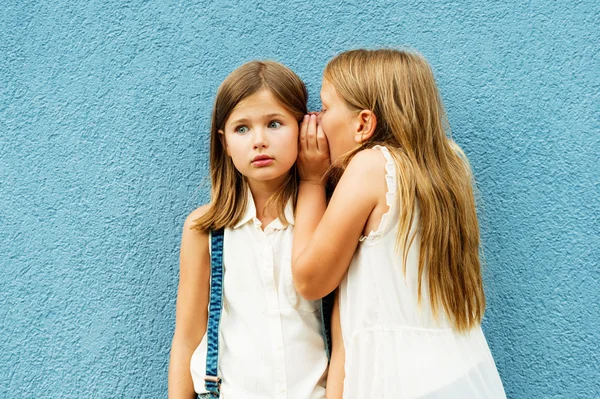 Dwie śliczne dziewczynki dzielące sekrety, stojące przed niebieską ścianą na podwórku szkolnego — Zdjęcie stockowe