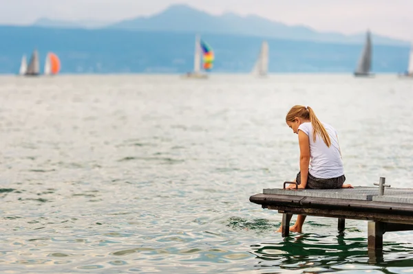 Linda niña descansando junto al lago, sentada en el muelle, salpicando agua con los pies, vista trasera — Foto de Stock