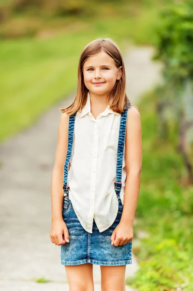 Outdoor portrait of a cute little girl wearing white shirt and denim skirt with suspenders — Stock Photo, Image