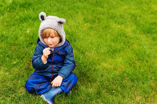 Criança bonito descansando ao ar livre, sentado em um gramado, jogando em um dia chuvoso, vestindo azul impermeável tudo-em-um terno — Fotografia de Stock