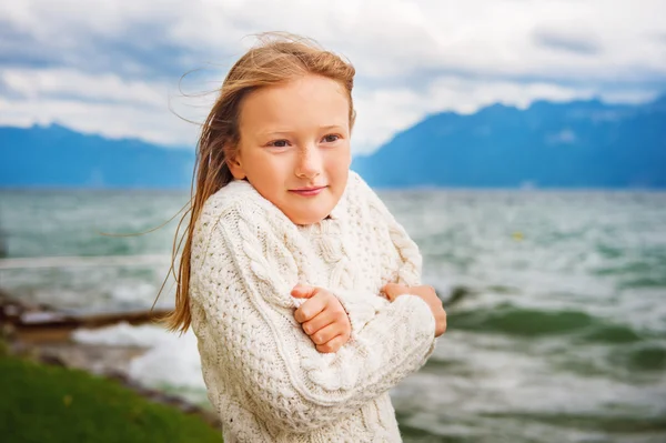 Милая маленькая девочка 8 лет играет на озере в очень ветреный день, одетая в теплый белый вязаный свитер — стоковое фото