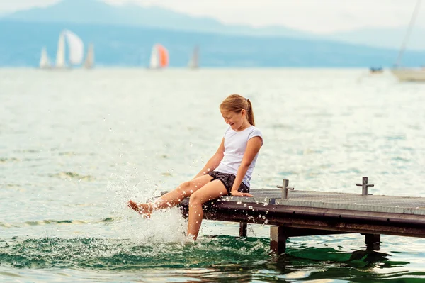 Śliczne małe dziecko dziewczyna odpoczynku nad jeziorem, siedząc na molo, rozpryskiwania wody z nogami — Zdjęcie stockowe