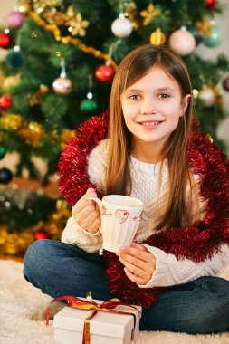 Noel ağacının yanında oturan küçük tatlı bir kızın portresi, beyaz bir kazak giymiş, elinde sıcak çikolatayla, kış tatilinde