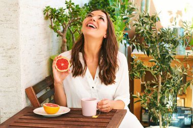 Genç mutlu kadın balkonda kahvaltı yapıyor, greyfurt ya da kırmızı portakal yiyor, masada bir fincan tişört yiyor.