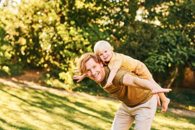 Mutlu genç baba yaz parkında oğluyla oynuyor, sırtına biniyor.