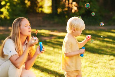 Mutlu genç anne ve sevimli çocuk yaz parkında sabun köpüğüyle oynuyorlar, mutlu bir çocukluk.