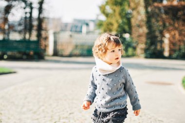 1-2 yaşında, gri mavi örgü kazak giyen, parkta oynayan sevimli bir kızın açık hava portresi.