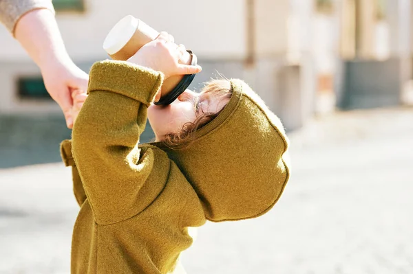 Komik çocuk dışarıda yürüyor, kağıt bardaktan içiyor, annesinin elini tutuyor.